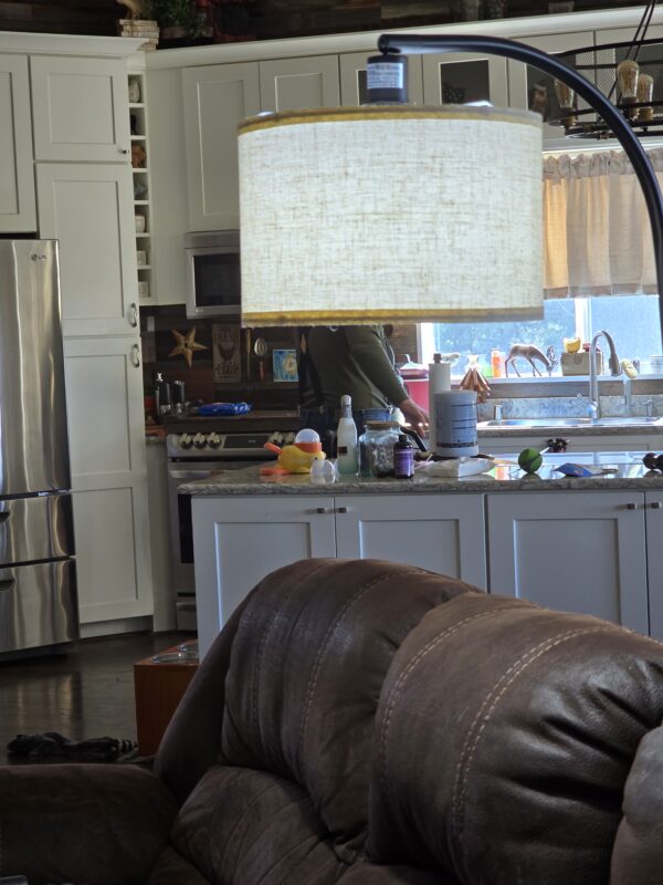 Bright kitchen with white cabinets, stainless fridge, and a large beige lampshade over the island; a person stands at the counter in the background.