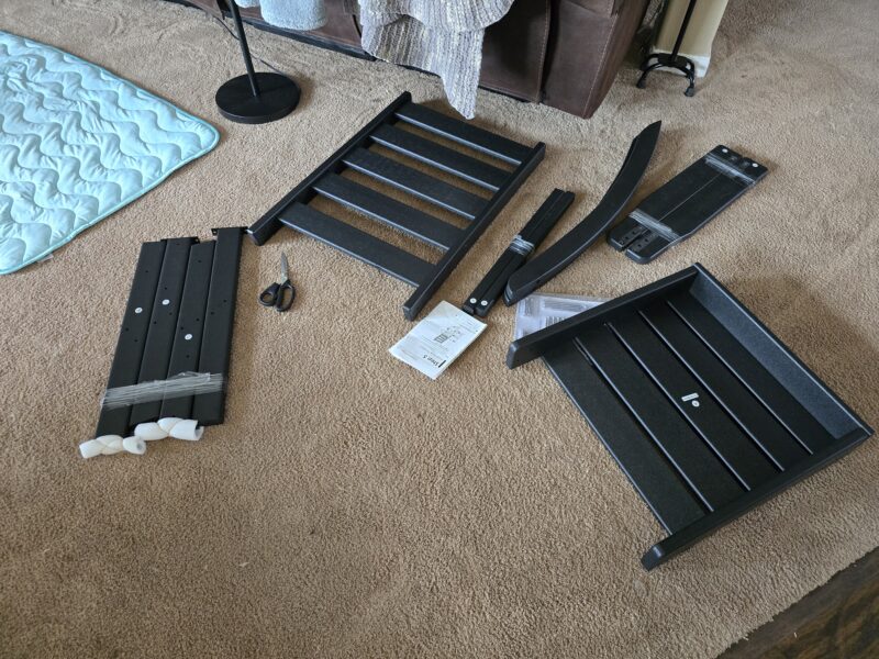 Disassembled Greenvines rocking chair frame parts on a carpet: metal slats, side rails, end rails, a pair of scissors, and an instruction manual 