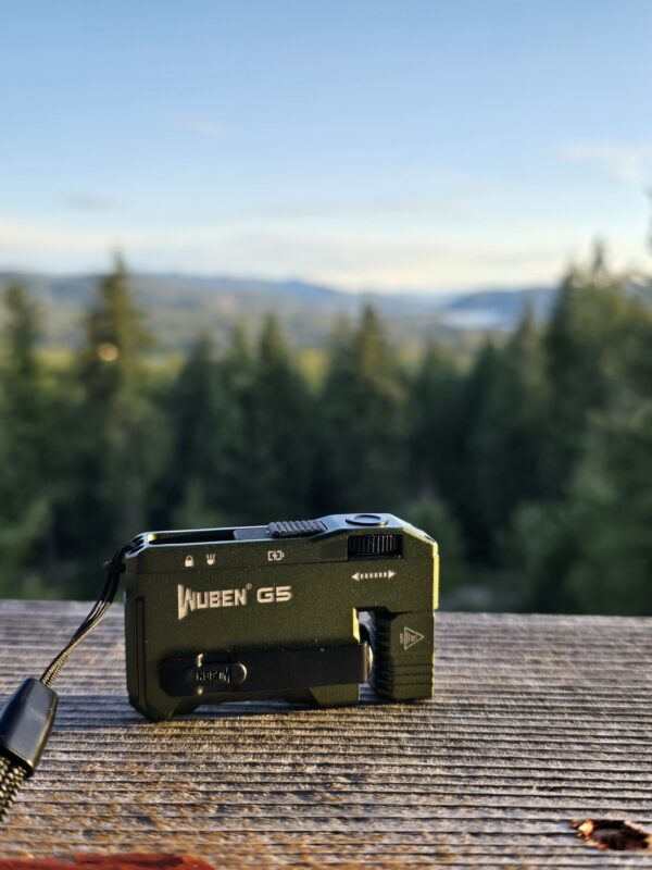 Black Wuben G5 flashlight resting on a weathered wooden surface with a blurred forest and distant mountains in the background.