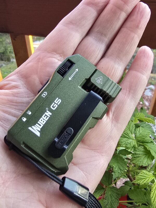 Close-up of a small green handheld utility knife or cutter held in an open palm, with a safety slider and textured grip, outdoors among plants.