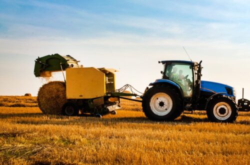 A side view of a blue farm tractor in a straw field towing a yellow round hay baler as it harvests straw for hay bales.