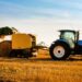 A side view of a blue farm tractor in a straw field towing a yellow round hay baler as it harvests straw for hay bales.
