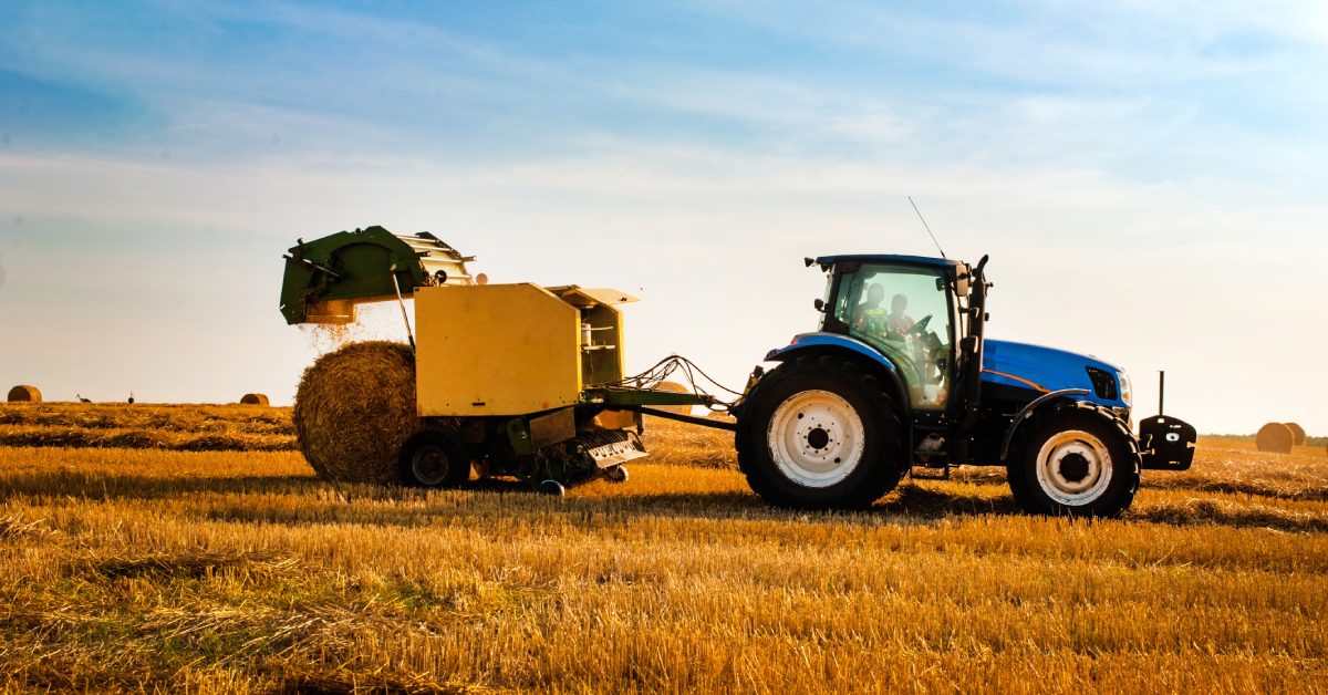 A side view of a blue farm tractor in a straw field towing a yellow round hay baler as it harvests straw for hay bales.
