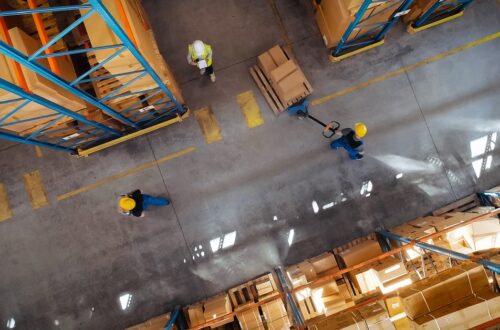 Top view of workers in warehouse aisle transporting stacked boxes on pallet jack between storage racks.