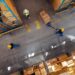 Top view of workers in warehouse aisle transporting stacked boxes on pallet jack between storage racks.