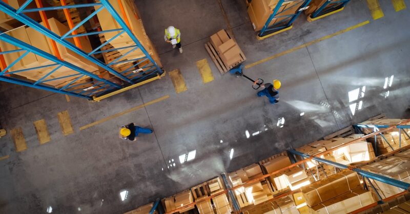 Top view of workers in warehouse aisle transporting stacked boxes on pallet jack between storage racks.