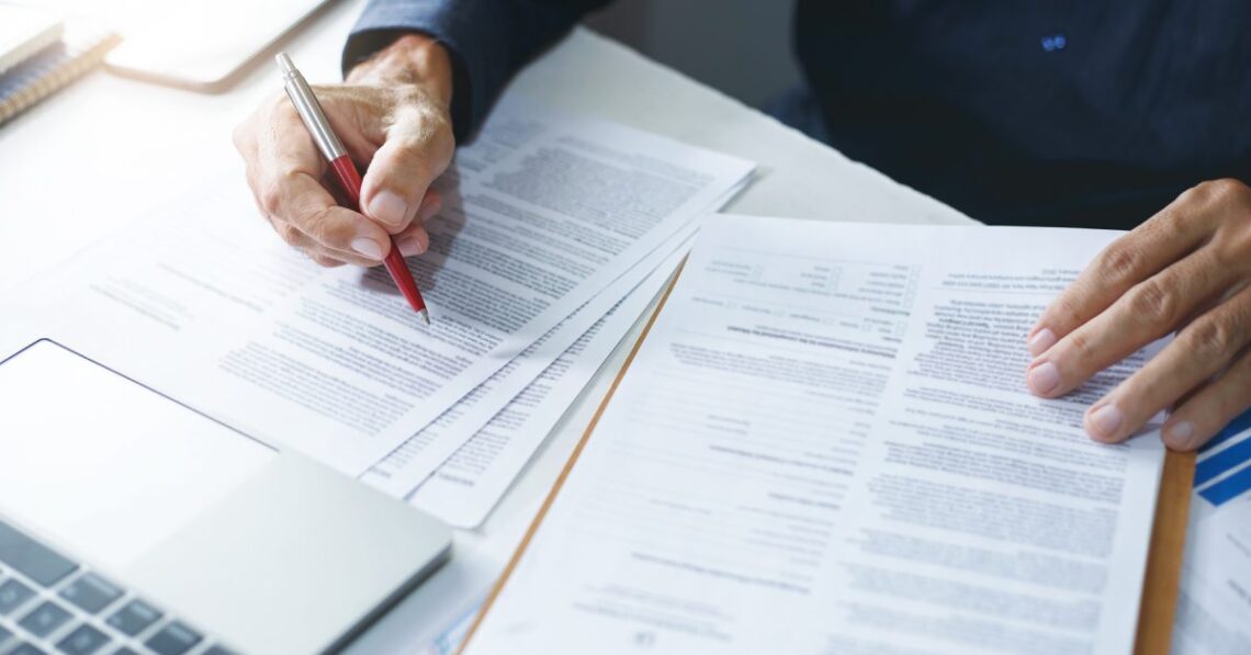 Close view of hands editing paperwork, using red pen to annotate pages near a laptop and documents on a clipboard.