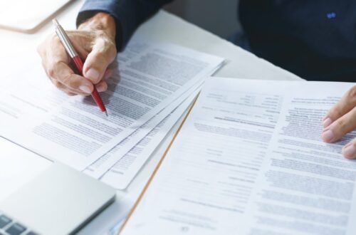 Close view of hands editing paperwork, using red pen to annotate pages near a laptop and documents on a clipboard.