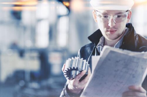 A factory engineer wearing a hard hat inspects a manufactured part in an industrial setting during a quality check.