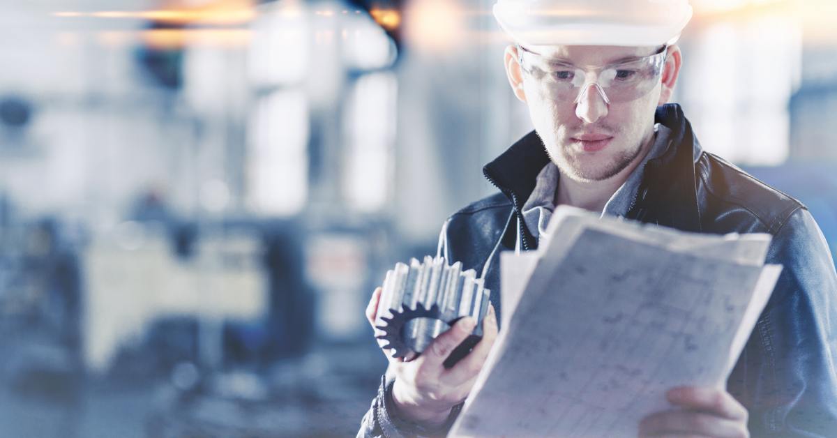 A factory engineer wearing a hard hat inspects a manufactured part in an industrial setting during a quality check.