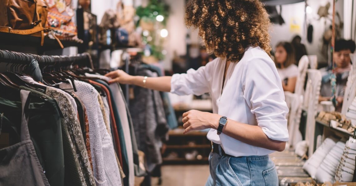 A woman is looking at a rack of clothing. The woman has voluminous dark hair and is wearing a white shirt.