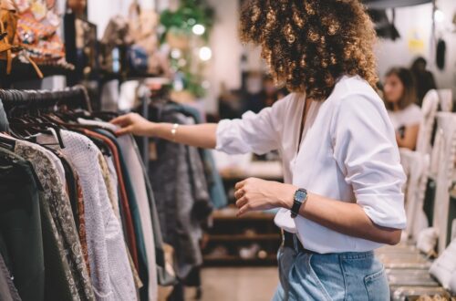 A woman is looking at a rack of clothing. The woman has voluminous dark hair and is wearing a white shirt.