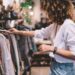A woman is looking at a rack of clothing. The woman has voluminous dark hair and is wearing a white shirt.