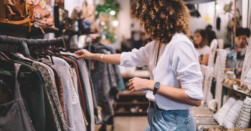 A woman is looking at a rack of clothing. The woman has voluminous dark hair and is wearing a white shirt.