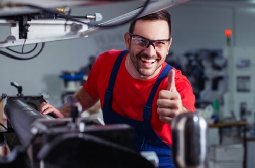 An industrial worker smiles and gives a thumbs-up while working on a machine in a factory. He wears safety glasses.