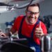 An industrial worker smiles and gives a thumbs-up while working on a machine in a factory. He wears safety glasses.