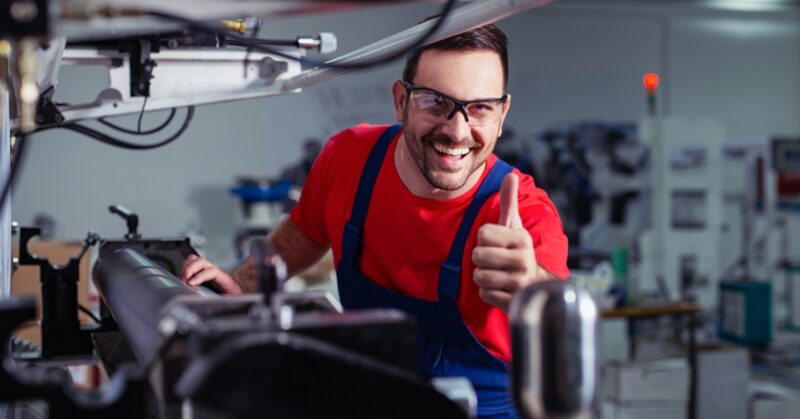 An industrial worker smiles and gives a thumbs-up while working on a machine in a factory. He wears safety glasses.