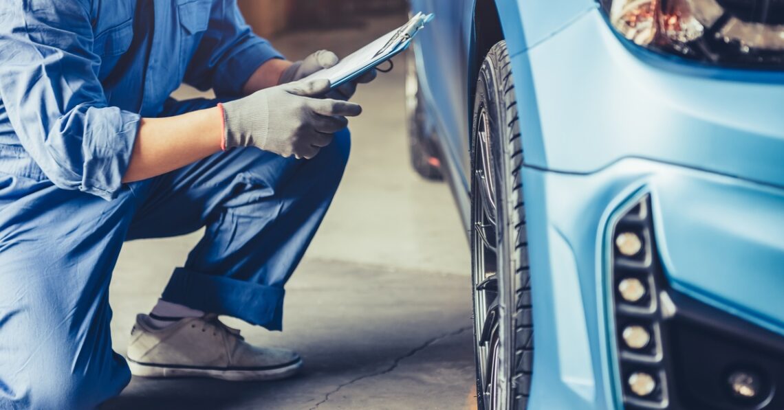 Mechanic kneeling beside car, inspecting front wheel while holding clipboard in automotive service garage.