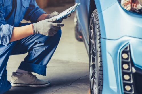 Mechanic kneeling beside car, inspecting front wheel while holding clipboard in automotive service garage.