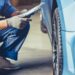 Mechanic kneeling beside car, inspecting front wheel while holding clipboard in automotive service garage.