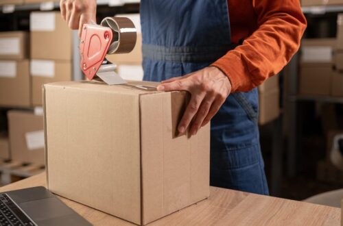 A person uses a tape dispenser to seal a cardboard box on a table, with stacked boxes visible in the background.