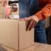 A person uses a tape dispenser to seal a cardboard box on a table, with stacked boxes visible in the background.
