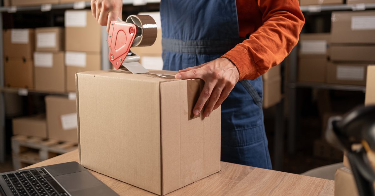 A person uses a tape dispenser to seal a cardboard box on a table, with stacked boxes visible in the background.