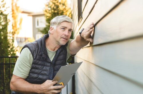 Man inspecting exterior house siding, touching wall while holding clipboard during home maintenance check.