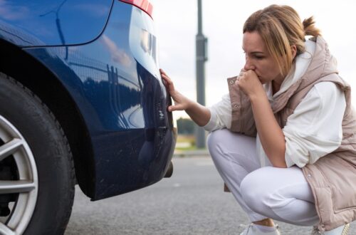 A woman looking at her back bumper after a minor accident. She is crouched behind the vehicle, her hand over her mouth.