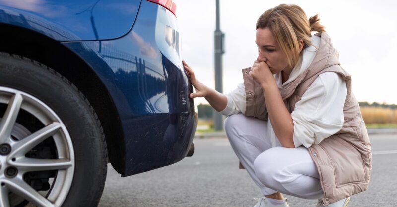 A woman looking at her back bumper after a minor accident. She is crouched behind the vehicle, her hand over her mouth.
