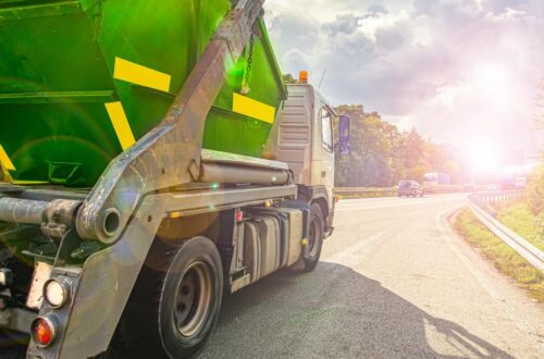 Large green dump trailer attached to a tow truck on a highway, with vehicles ahead and bright sun glare in the distance.