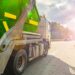 Large green dump trailer attached to a tow truck on a highway, with vehicles ahead and bright sun glare in the distance.