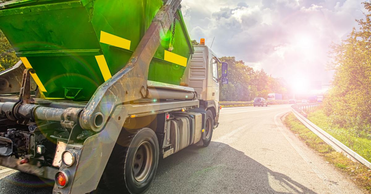 Large green dump trailer attached to a tow truck on a highway, with vehicles ahead and bright sun glare in the distance.