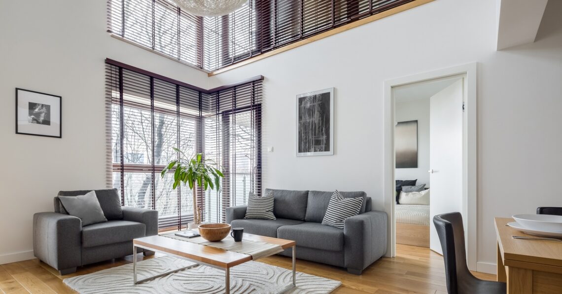 Modern living room with gray sofa and armchair, large windows with blinds, coffee table and rug in bright interior.