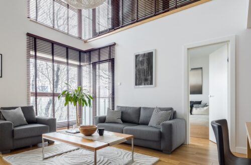 Modern living room with gray sofa and armchair, large windows with blinds, coffee table and rug in bright interior.