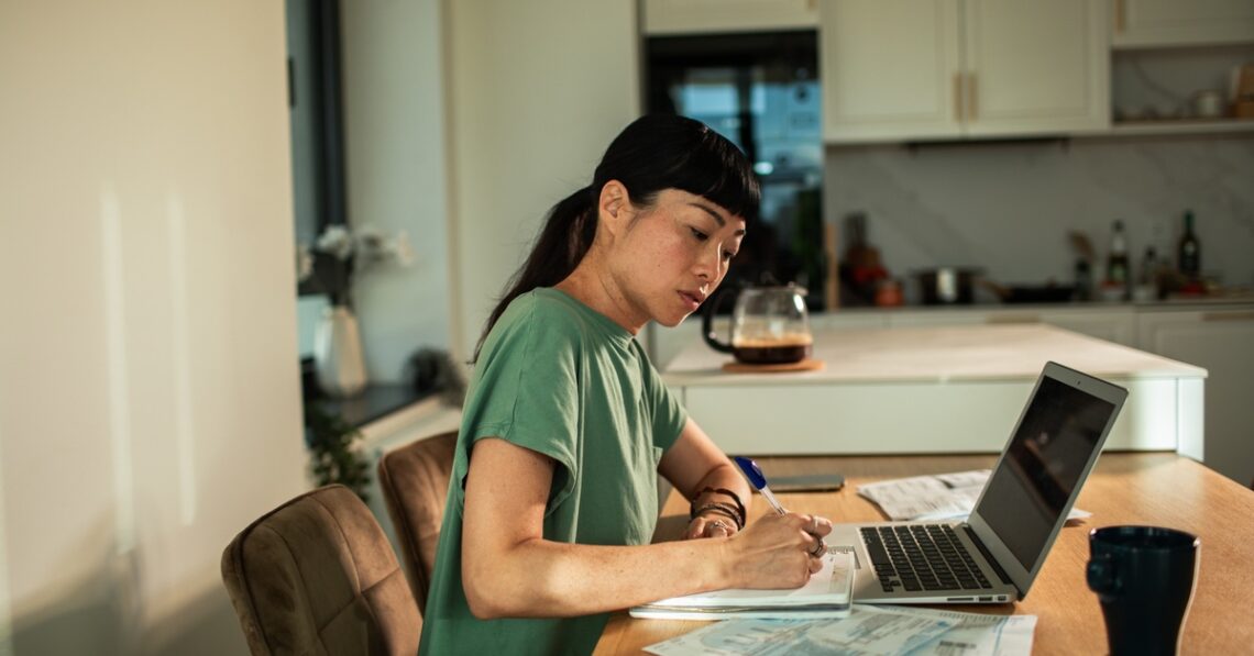 A young woman sits at her dining room table alone as she uses a laptop to track finances, writing things on paper.