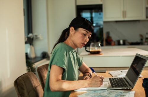 A young woman sits at her dining room table alone as she uses a laptop to track finances, writing things on paper.