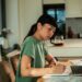 A young woman sits at her dining room table alone as she uses a laptop to track finances, writing things on paper.