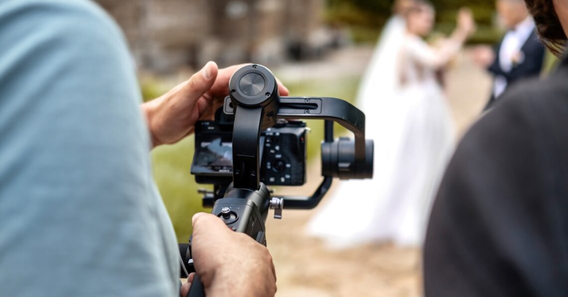 A person aiming a camera on a swivel mount toward a bride and groom. They're using both hands to hold the camera steady.