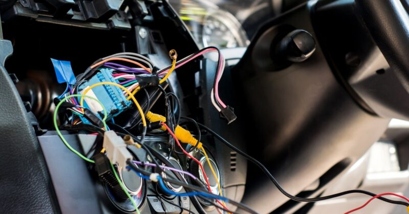 A car interior during the daytime. A panel is popped out of the dashboard to reveal colorful wires sticking out.