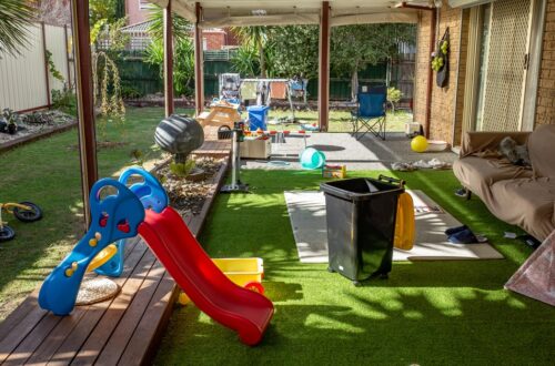 A covered backyard patio contains toys, a couch, and scattered clutter as the family space spills into the yard.