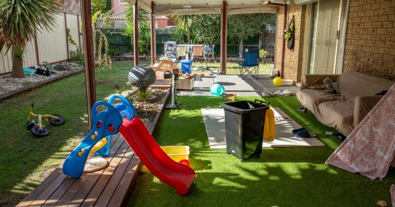 A covered backyard patio contains toys, a couch, and scattered clutter as the family space spills into the yard.