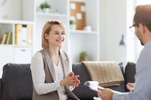 A smiling blonde woman sits on a gray couch during a therapy session and speaks with an older male therapist holding notes.