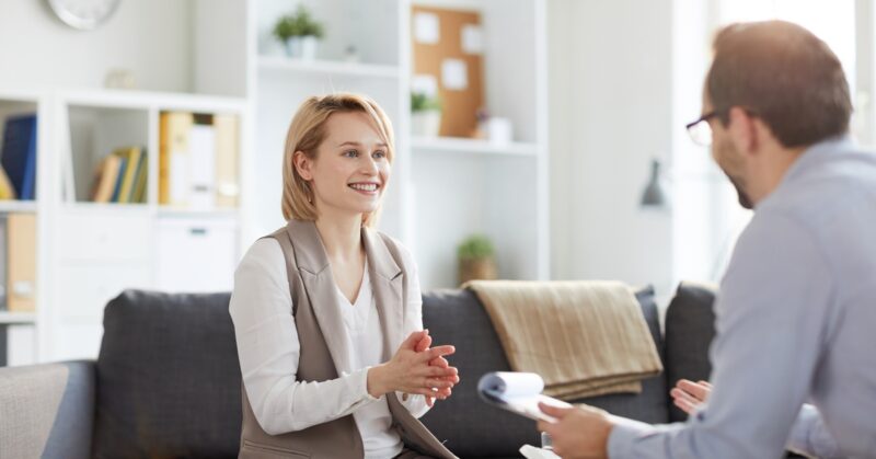 A smiling blonde woman sits on a gray couch during a therapy session and speaks with an older male therapist holding notes.
