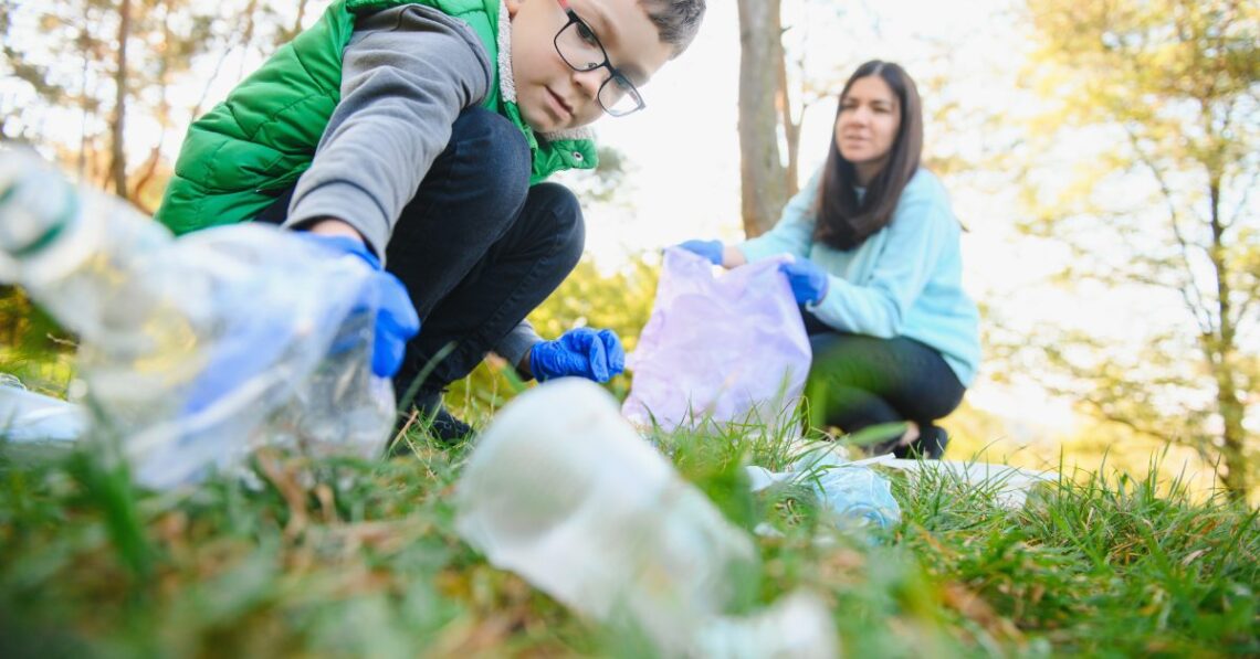A child crouching down to pick up a plastic bottle while wearing blue gloves as a parent holds a trash bag for them.