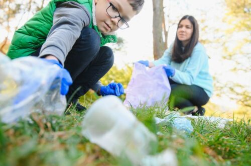 A child crouching down to pick up a plastic bottle while wearing blue gloves as a parent holds a trash bag for them.