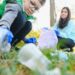 A child crouching down to pick up a plastic bottle while wearing blue gloves as a parent holds a trash bag for them.