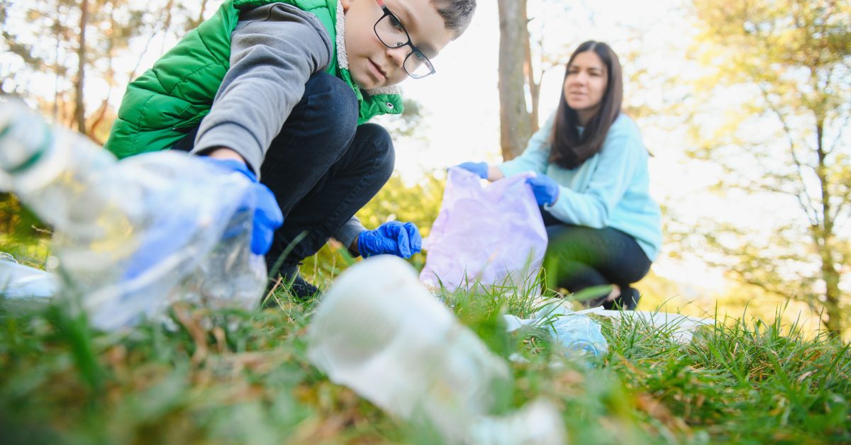 A child crouching down to pick up a plastic bottle while wearing blue gloves as a parent holds a trash bag for them.