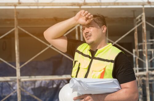Construction worker in a neon safety vest shields his eyes from the sun while holding blueprints and a white hard hat on a scaffolding site.