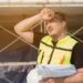 Construction worker in a neon safety vest shields his eyes from the sun while holding blueprints and a white hard hat on a scaffolding site.